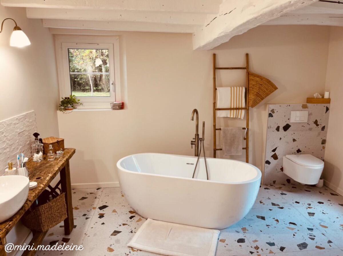 Terrazzo multicouleur avec éclats de tons gris, noir, blanc, dimension 80x80 cm dans une salle de bain beige avec baignoire ovale, meuble en bois, échelle décorative et textiles écrus