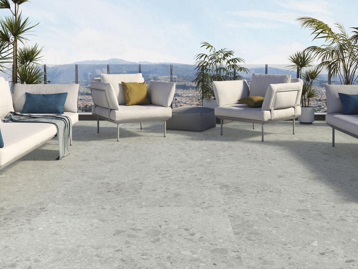 Carrelage Terrazzo gris clair nuances de blanc 60x60 cm sur une terrasse mobilier moderne coussins bleus et jaunes verdure ville en arrière-plan