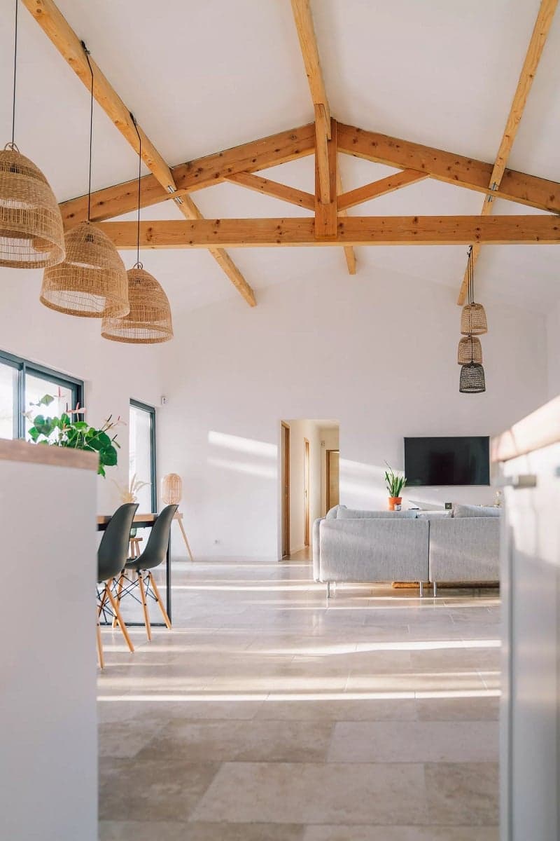 Carrelage effet pierre beige dans un salon lumineux avec poutres en bois, mobilier moderne, plantes vertes et éclairage naturel