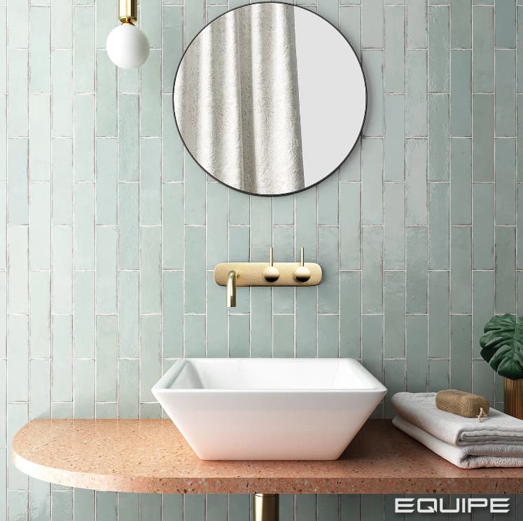 Carrelage Zellige vert clair uni sur mur de salle de bain épurée avec lavabo blanc sur plan de travail terrazzo beige et accessoires dorés
