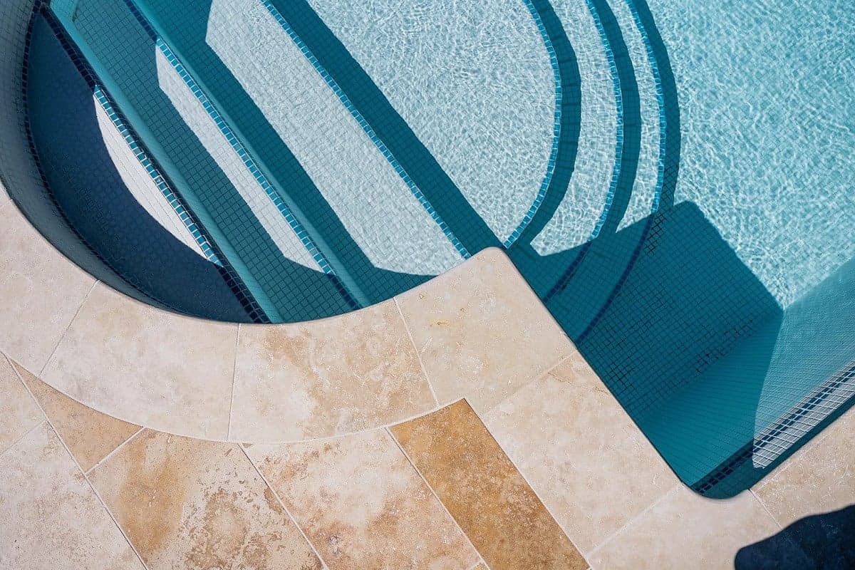 Carrelage effet pierre beige sans motifs sur bord de piscine aux eaux turquoise clair entourée de bleu foncé