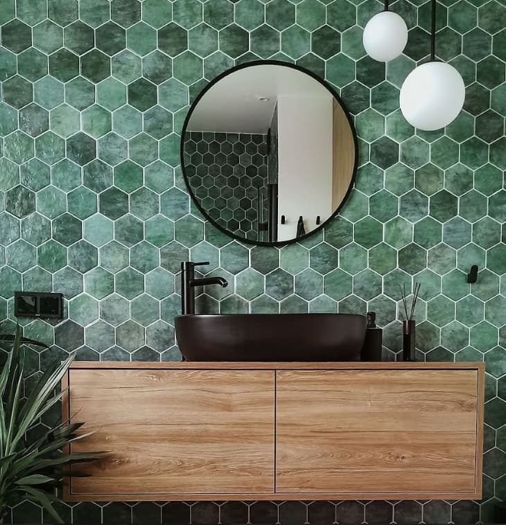 Zellige vert émeraude brillant sur mur salle de bains avec meuble bois marron, vasque noire, miroir rond, luminaires suspendus blancs