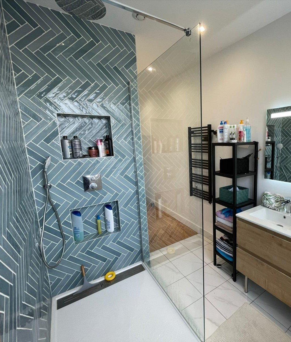 Zellige bleu en chevron 5X25 sur un mur de salle de bain blanc, mobilier bois, étagère noire, accessoires de douche et baignoire