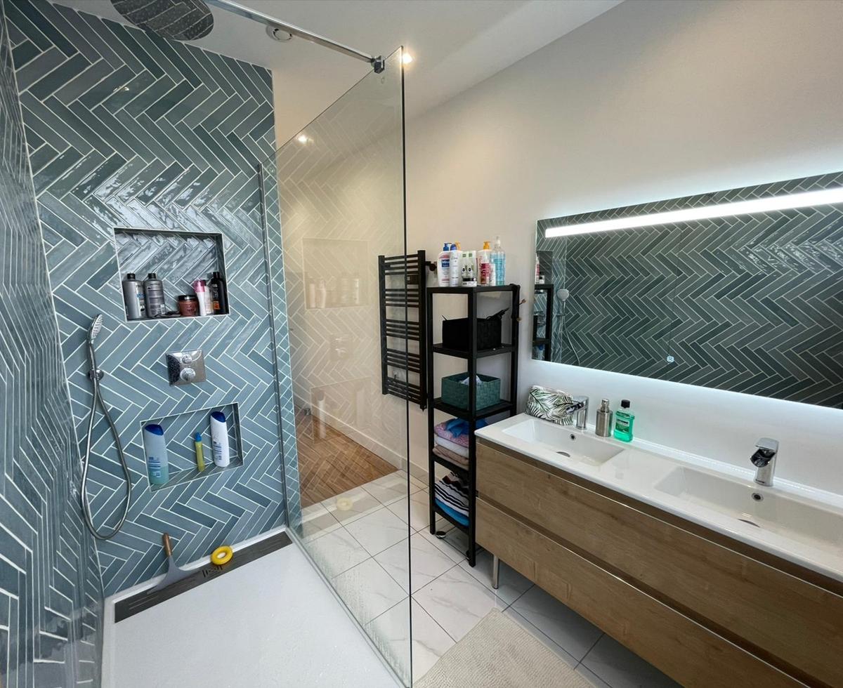 Zellige bleu en chevron 5X25 sur un mur de salle de bain blanc, mobilier bois, étagère noire, accessoires de douche et baignoire
