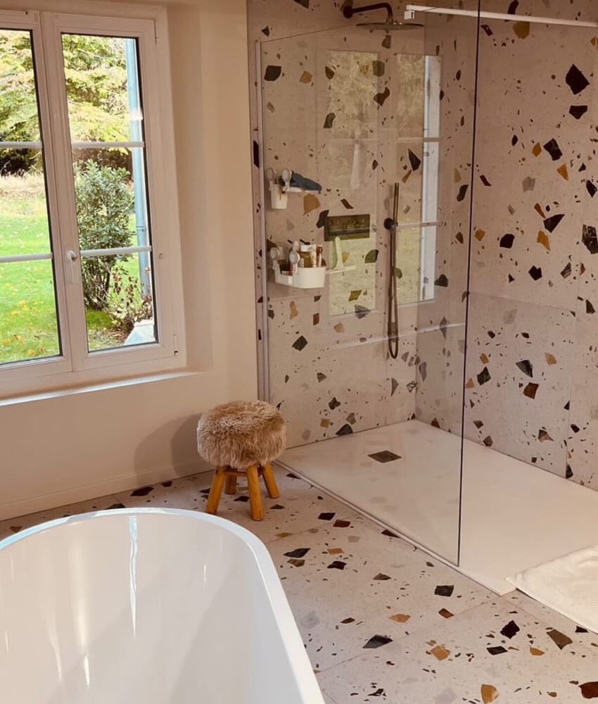 Carrelage Terrazzo blanc avec éclats multicolores 60x60 cm dans une salle de bain beige baignoire blanche et tabouret en bois