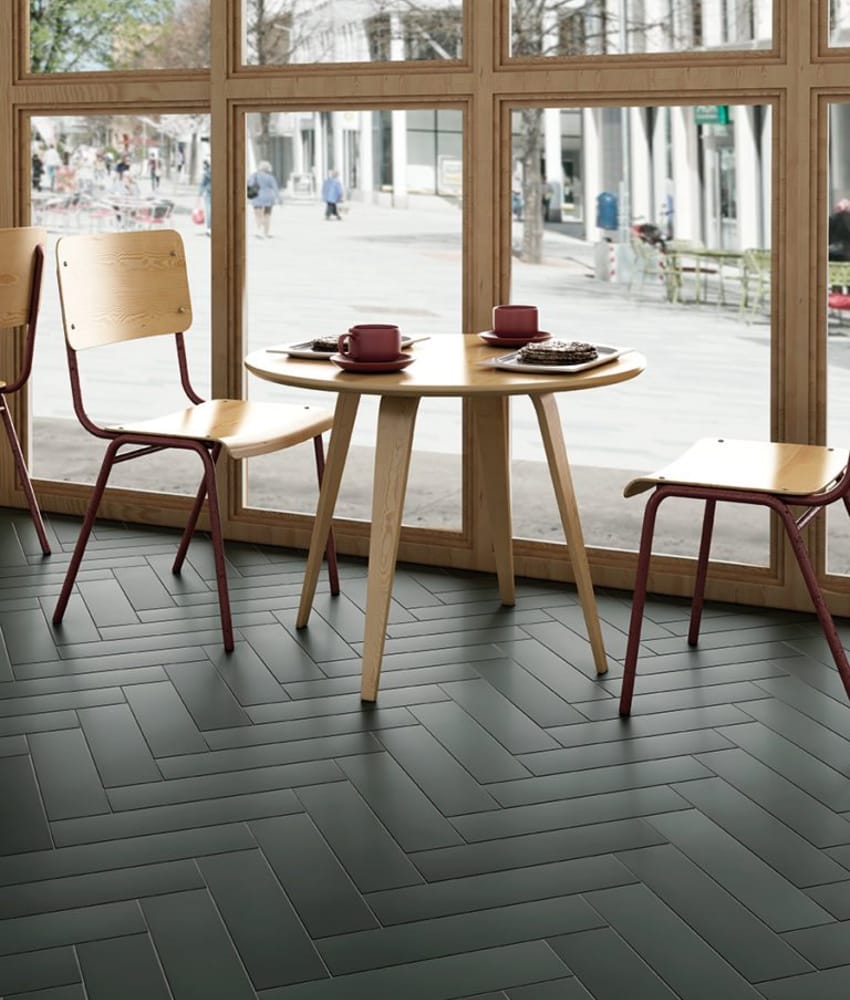 Carrelage uni vert foncé sur le sol dans un café avec tables en bois, chaises rouges et grandes fenêtres donnant sur la rue